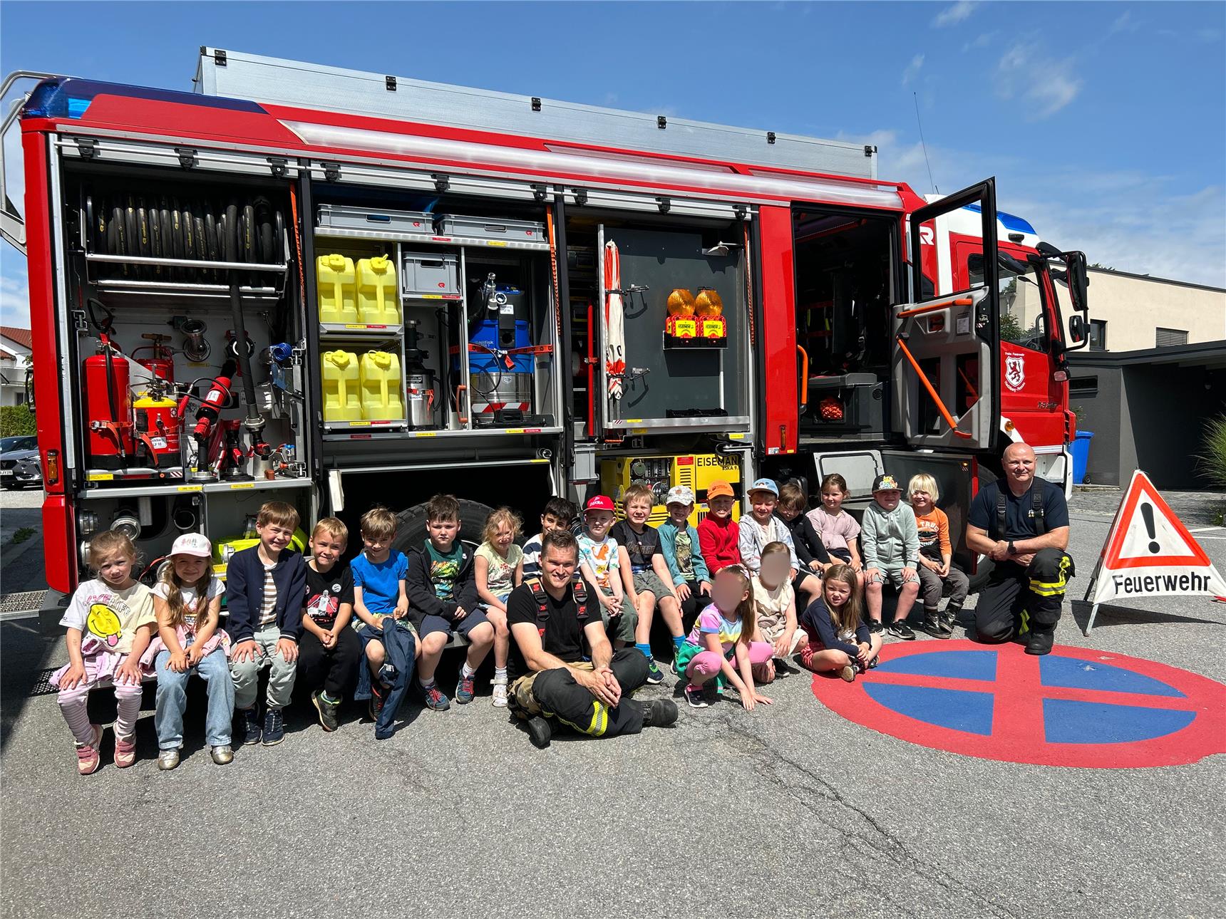 Eine Gruppe Vorschulkinder sitzt lachend vor einem geöffneten Feuerwehrauto mit zwei Feuerwehrmännern. Das Feuerwehrauto zeigt verschiedene Ausrüstungen wie Wasserschläuche, Kanister und Werkzeuge. Rechts steht ein Warndreieck mit der Aufschrift „Feuerwehr“.