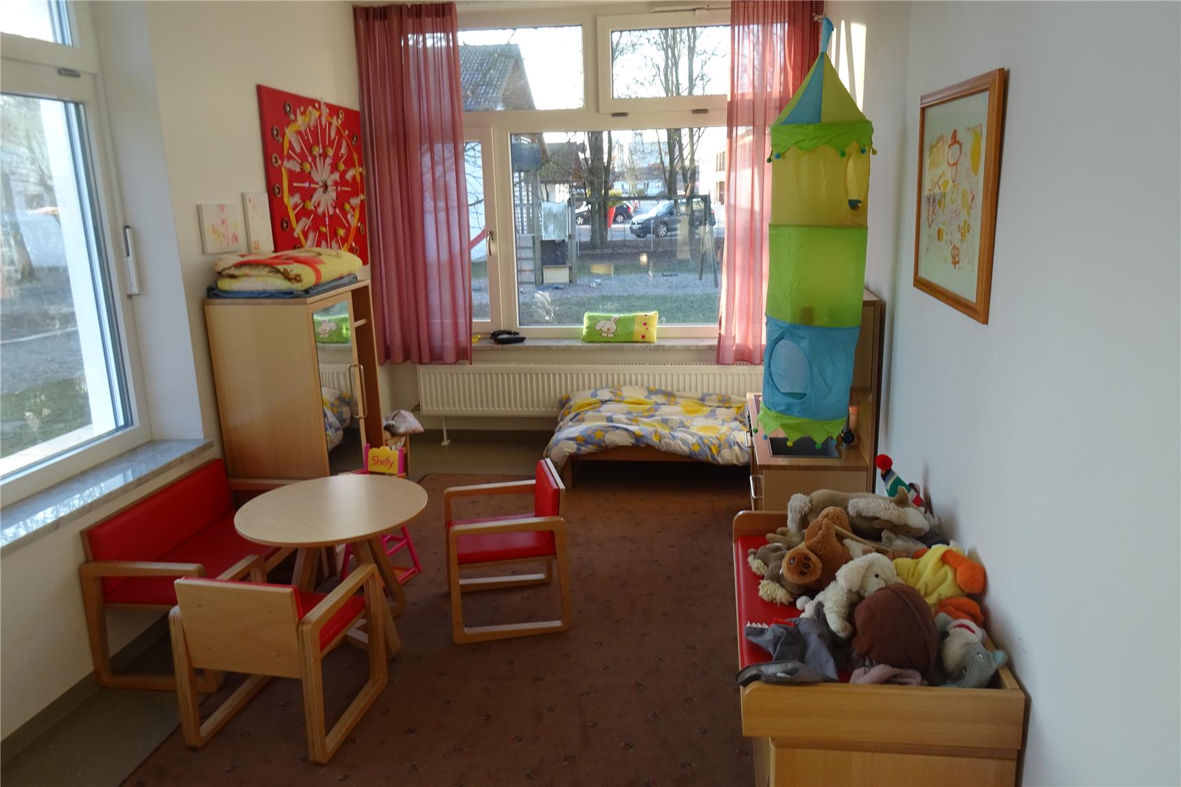 Gemütlich eingerichtetes Kinderzimmer mit kleinen Holzmöbeln, Kuscheltieren und einem bunten Hochbett-Turm. Große Fenster mit rosa Vorhängen lassen viel Tageslicht herein.