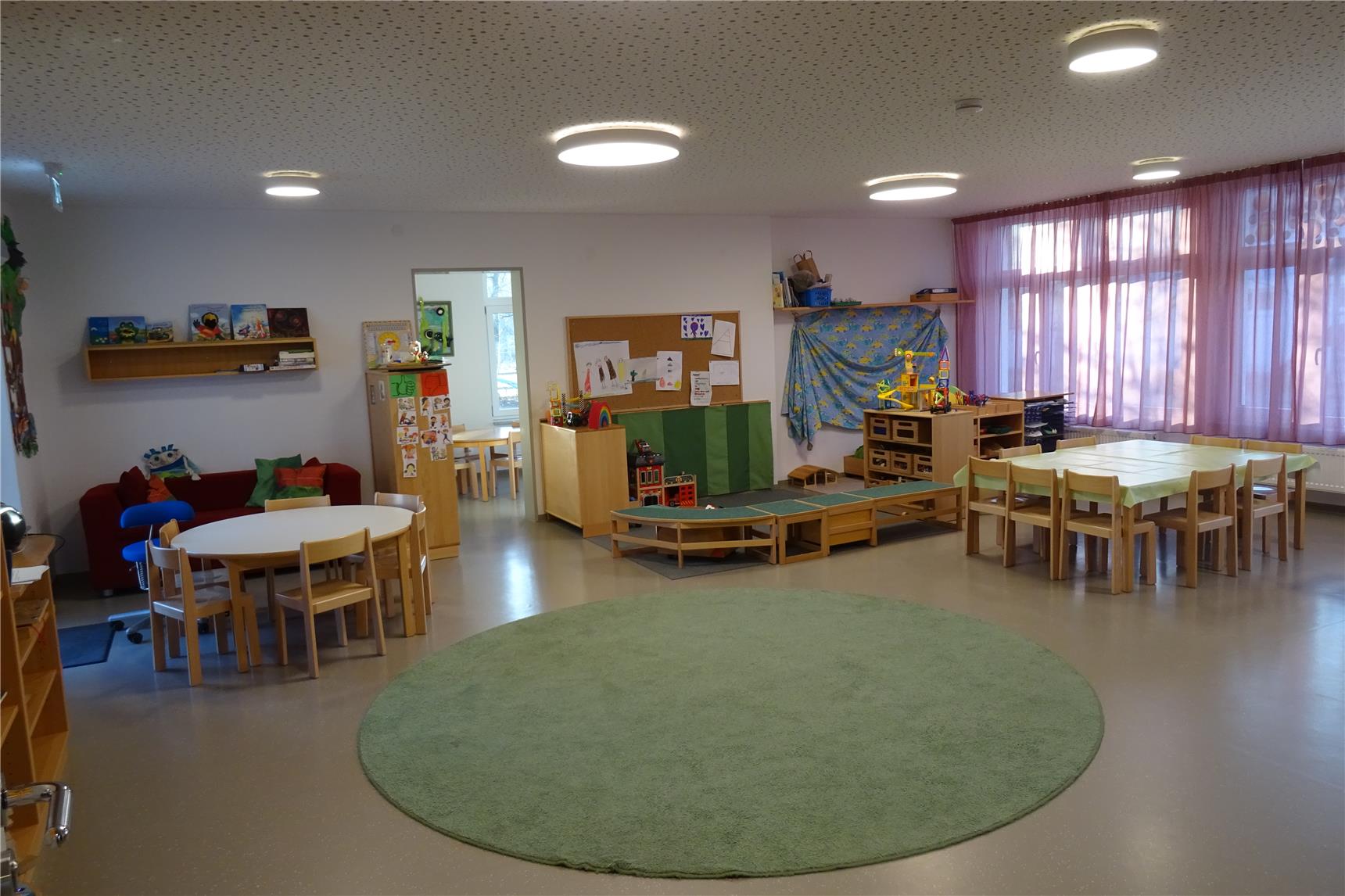 Heller Gruppenraum in einer Kita mit mehreren Holztischen, Spiel- und Leseecken sowie einem großen grünen Teppich in der Mitte. An den Wänden sind Kinderzeichnungen und Regale mit Spielmaterialien zu sehen.