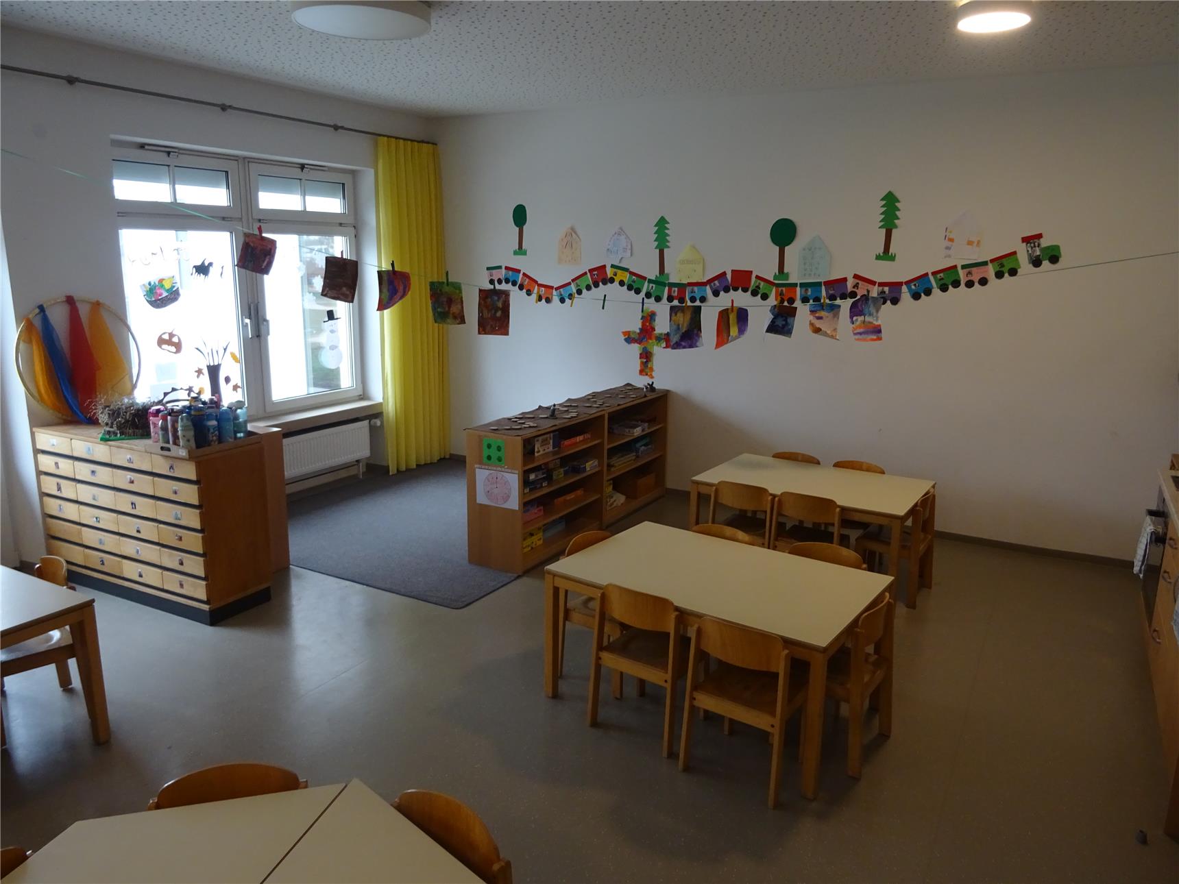 Heller Kindergartenraum mit kleinen Holztischen und Kinderstühlen, an der Wand hängt eine bunte Girlande aus gebastelten Zügen und Bildern. Auf der linken Seite stehen Schubladenschränke mit Bastelmaterialien und bemalten Flaschen.