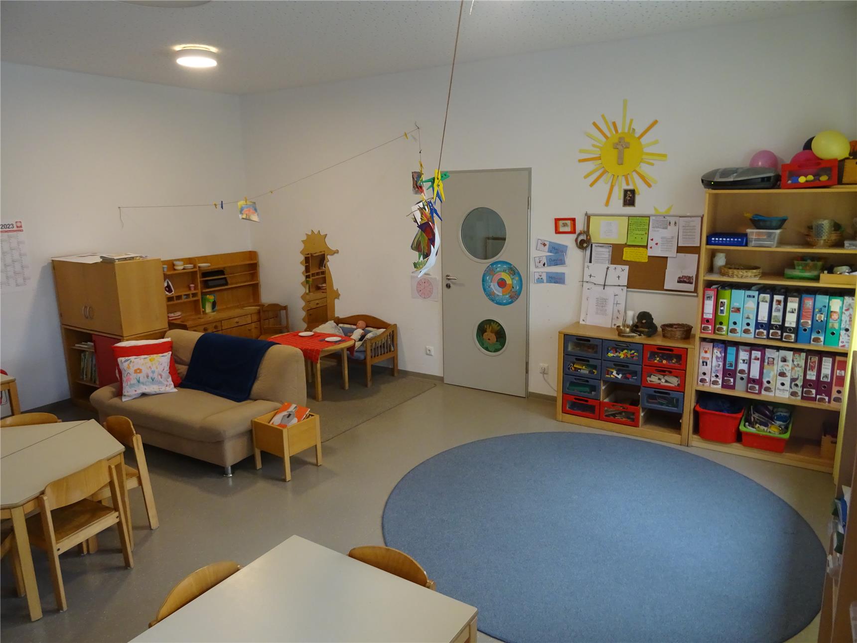 Gemütlicher Kindergartenraum mit Spielsofa, kleinem Bett und Bastelbereich, dekoriert mit Kinderzeichnungen und bunten Ordnern. An der Wand hängt eine gelbe Sonne mit Kreuz, daneben eine Pinnwand mit Infos und Kunstwerken.