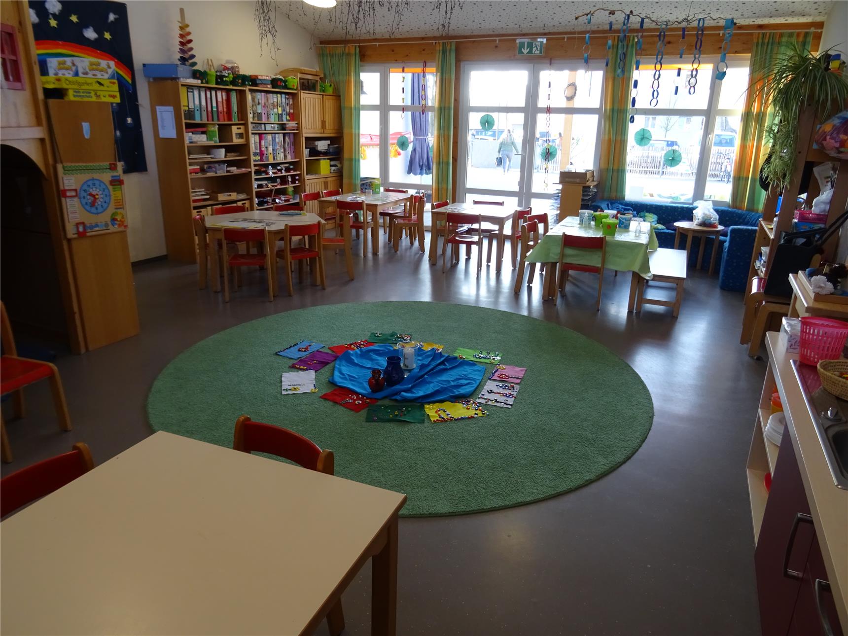 Ein Kindergartenraum mit Tischen und Stühlen und einem Spielteppich.