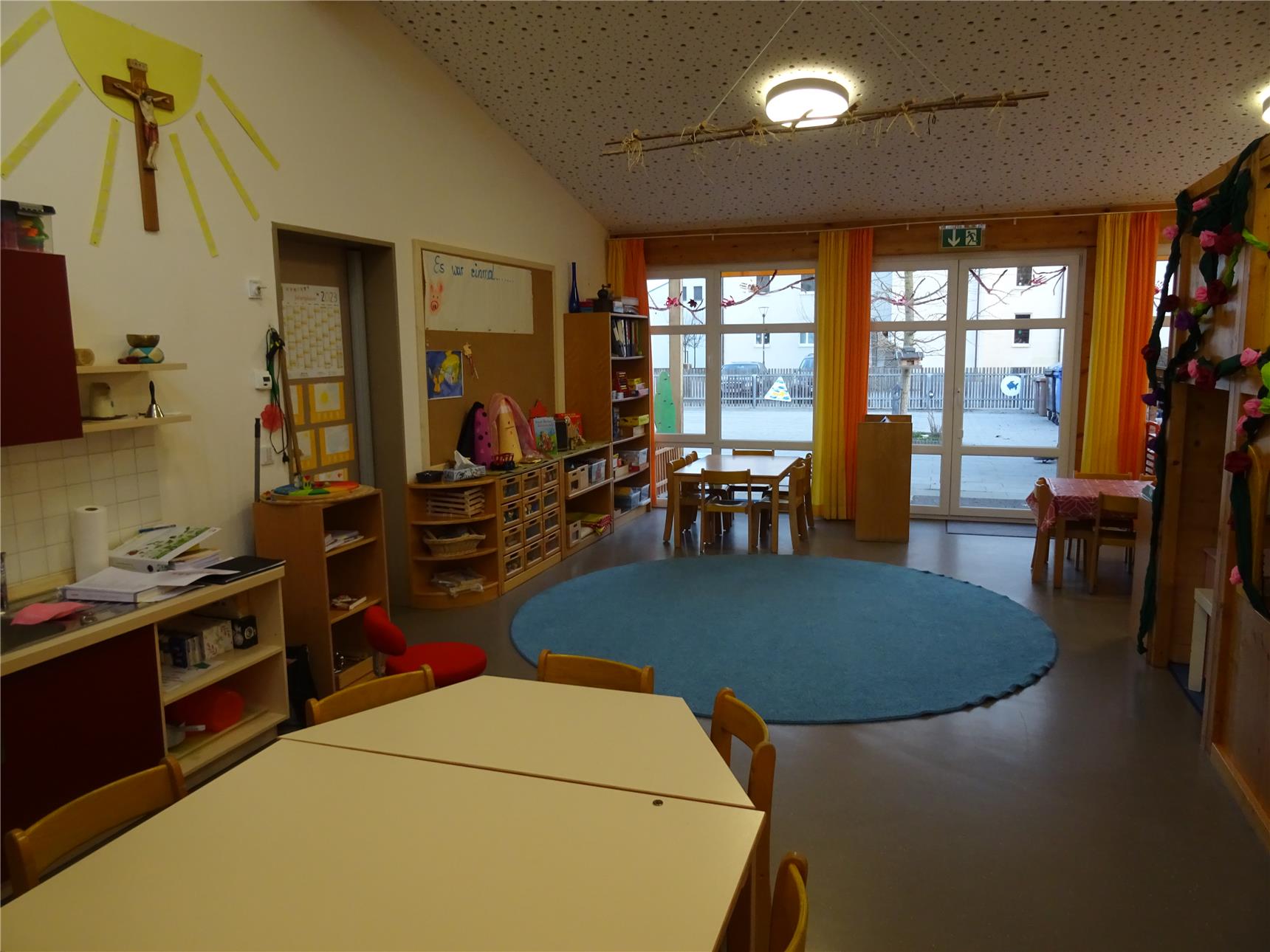 Ein großer, freundlicher Kindergartenraum mit mehreren Tischen und Stühlen. In der Raummitte liegt ein großer Spielteppich