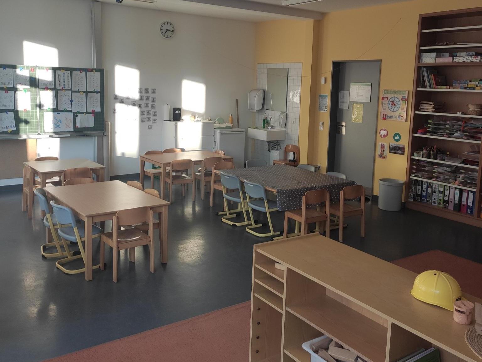 Heller Raum mit mehreren kleinen Holztischen und bunten Kinderstühlen. An der Wand hängen Kinderzeichnungen und im Hintergrund steht ein Waschbecken mit Seifenspender.