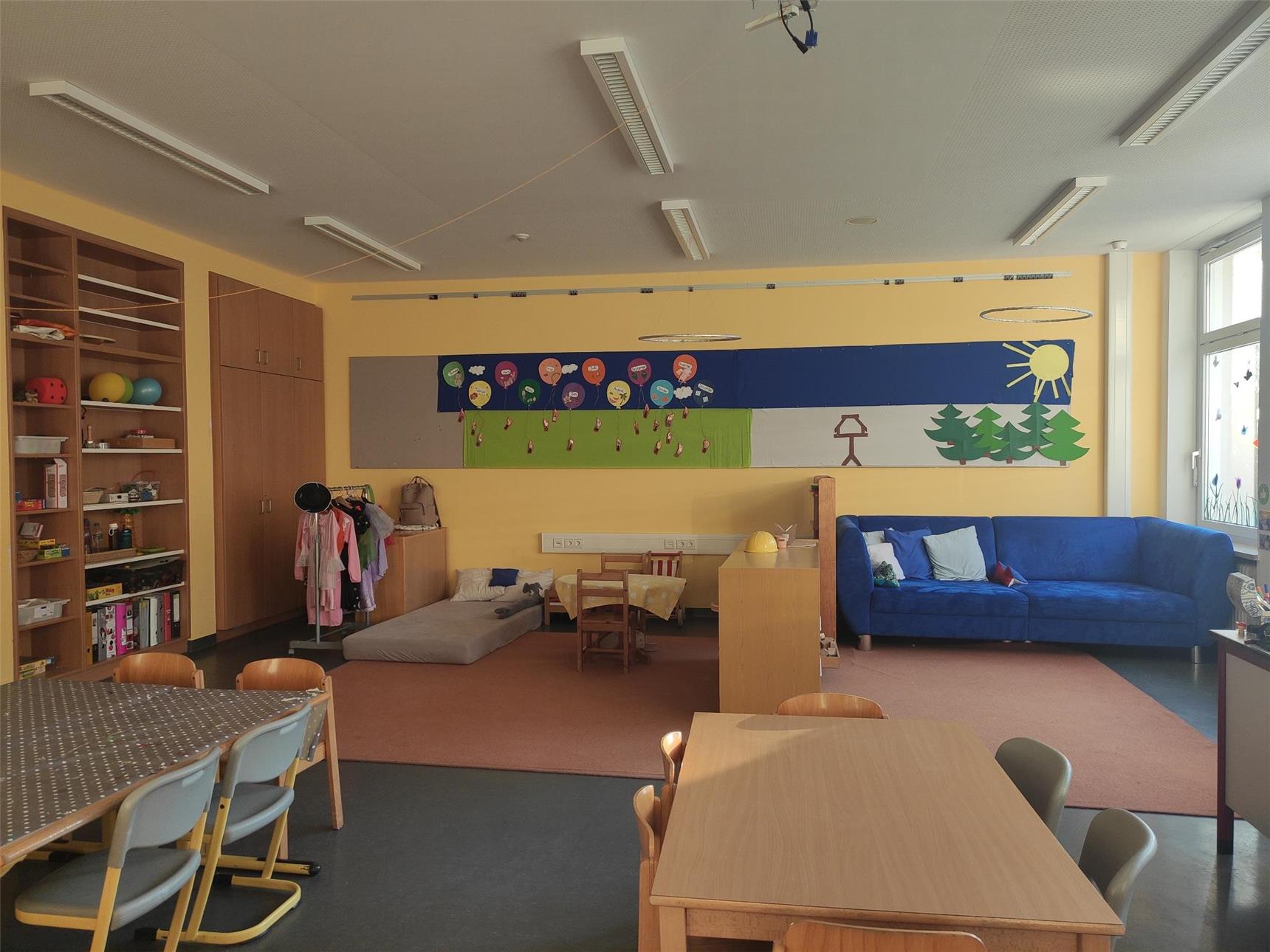 Gemütlicher Kindergartenraum mit Spielecke, blauer Couch und Bastelbereich. An der Wand hängt eine bunte Collage mit Bäumen, Sonne und Heißluftballons.