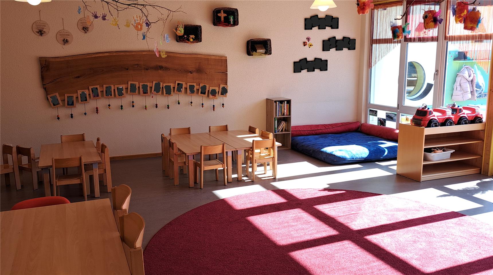 Heller und einladender Kindergartenraum mit kleinen Holztischen, roten Spielteppichen und einer gemütlichen Leseecke mit Kissen. An den Wänden hängen Kinderbasteleien und gerahmte Bilder.