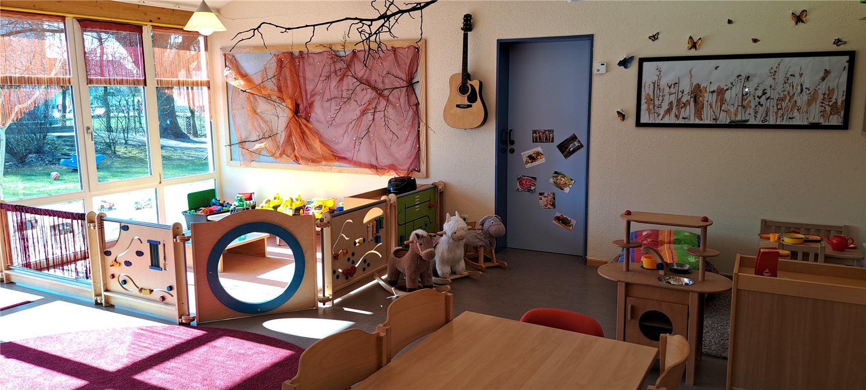 Spielbereich im Kindergarten mit bunter Spielküche, Stofftieren und Lernspielwänden. Große Fenster lassen viel Tageslicht herein, dahinter Blick auf den Garten.