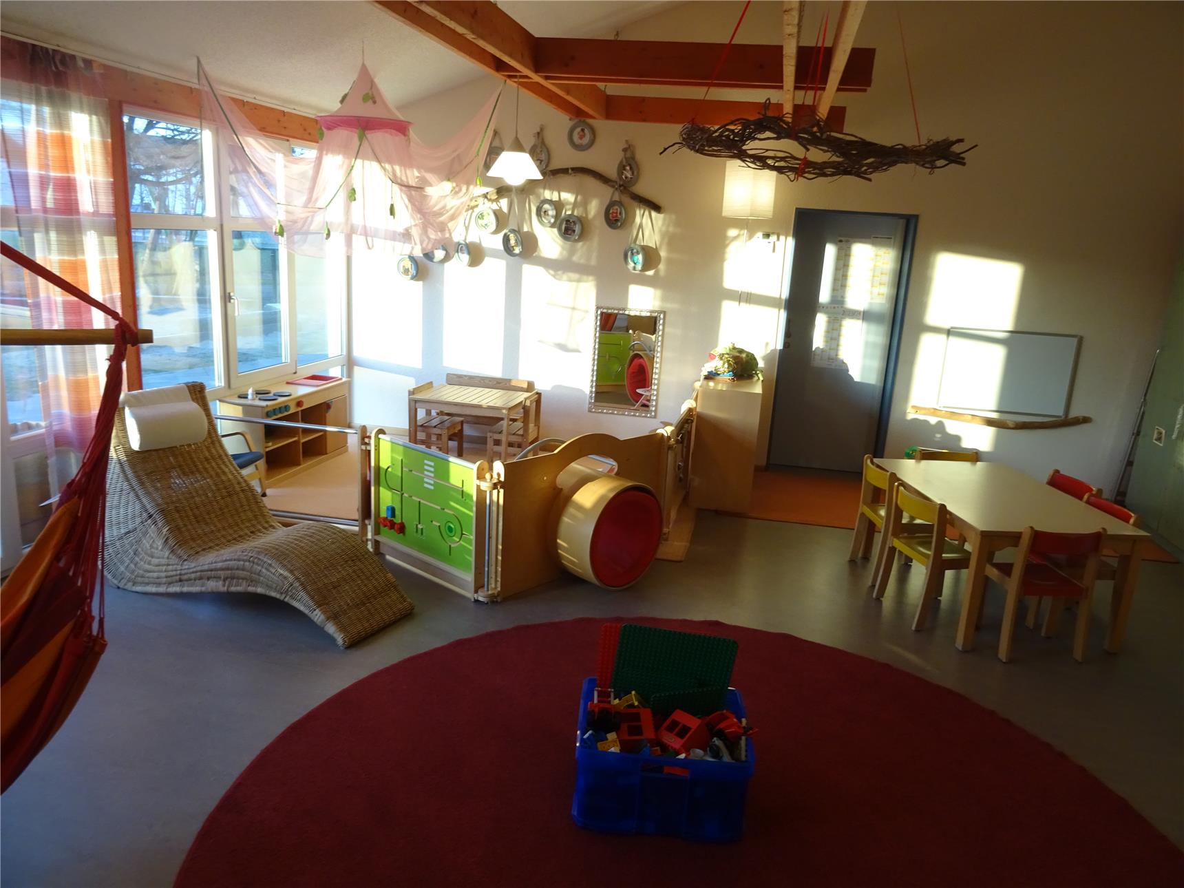 Gemütlicher Kindergartenraum mit Kletterröhre, Spielbereich und Basteltisch. Eine bunte Hängematte und ein Baldachin schaffen eine wohnliche Atmosphäre.