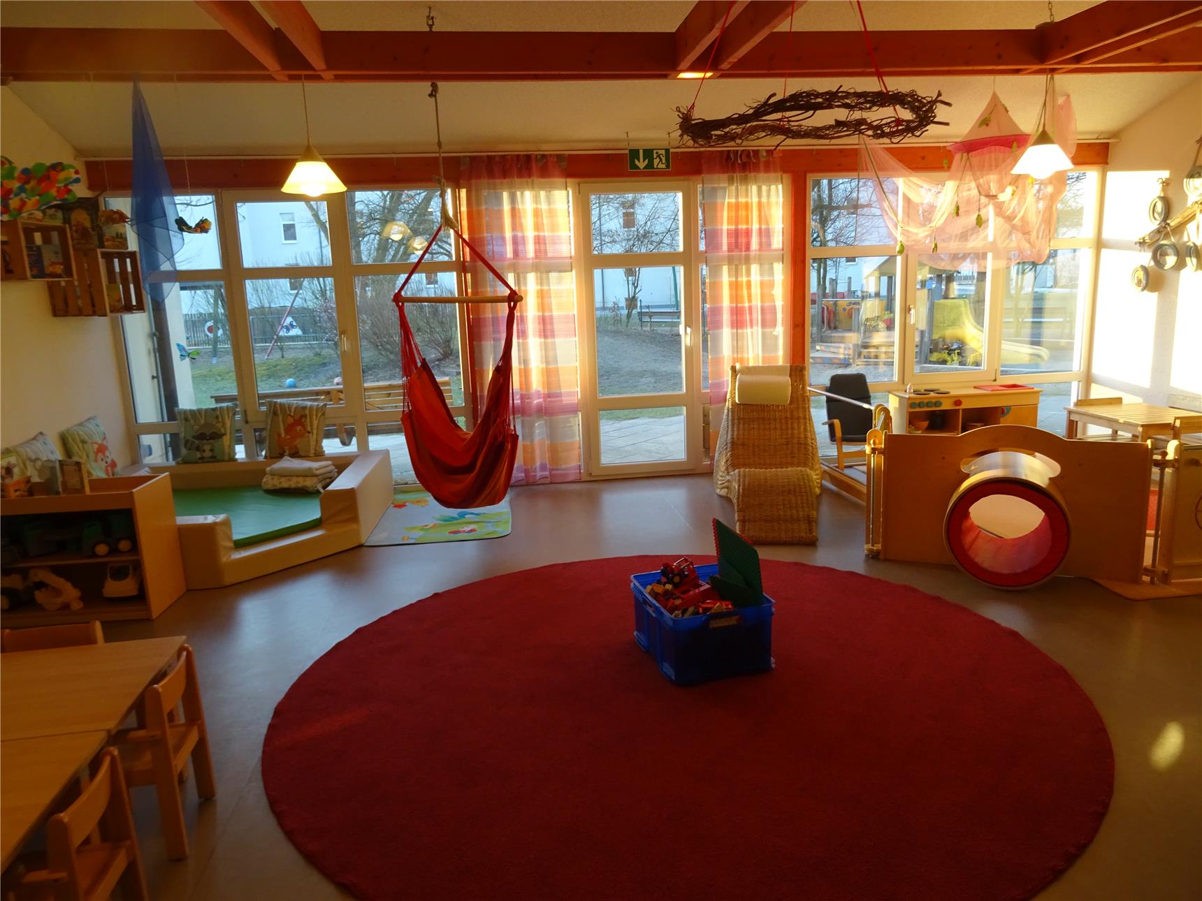 Kindergartenraum mit rotem Teppichkreis, Hängesessel und Spielröhre in gemütlicher Abendsonne. Kuschelecke mit bunten Kissen und Fenstern mit Blick nach draußen.