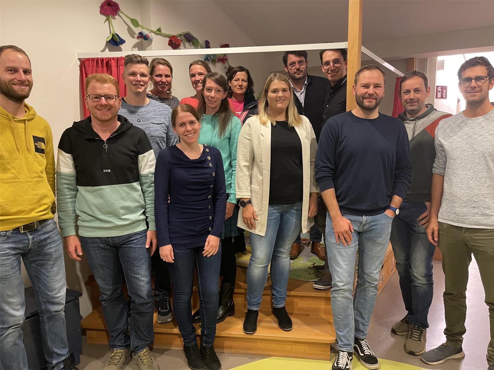 Gruppenfoto von 14 lächelnden Erwachsenen, die in einem Raum auf einer kleinen Treppe und davor stehen. Im Hintergrund sind bunte Papierblumen an der Wand dekoriert.        ChatGPT fragen