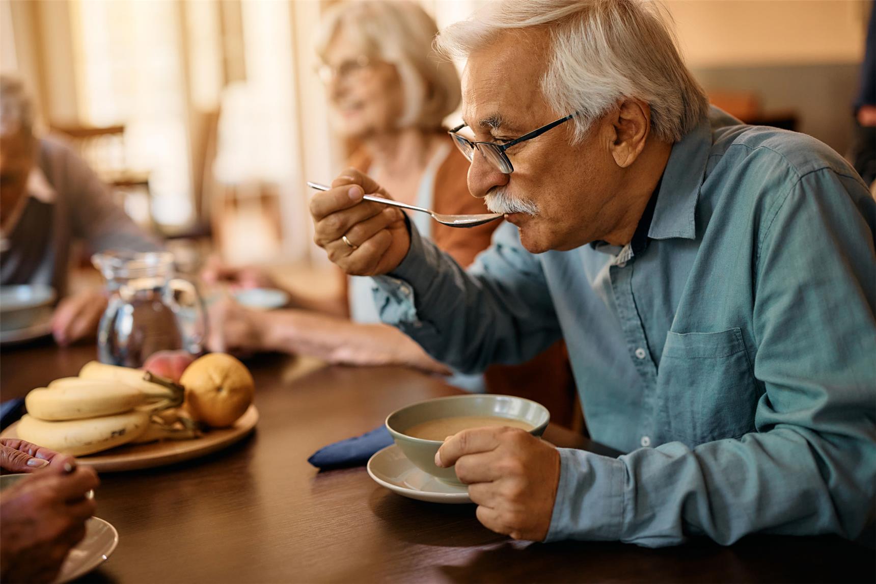 Ein älterer Mann und eine lächelnde ältere Frau sitzen am Esstisch. Sie unterhalten sich beim Essen mit weiteren Personen, die am Bildrand zu erkennen sind.
