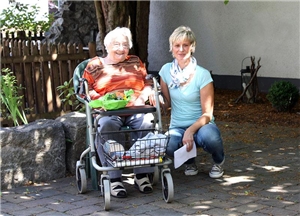 Seniorin im Rollstuhl sitzt im Garten, neben ihr kniet eine junge Frau.
