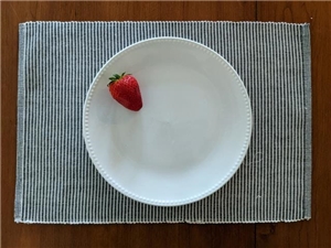 Ein weißer Teller steht auf einem gestreiften Stoff-Platzset, auf dem Teller liegt eine einzelne rote Erdbeere