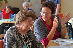 Eine Ehrenamtliche klatscht in die Hände, neben ihr sitzt eine lachende Seniorin.