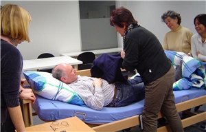 Eine Kursleiterin steht an einem Bett, in dem ein Mann liegt, die Teilnehmer schauen zu