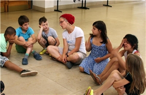 Das Foto zeigt eine Gruppe Kinder mit einer Betreuerin, die in einem großen Raum auf dem Boden sitzen.