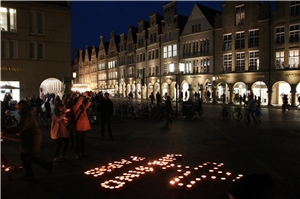 Das Foto zeigt brennende Kerzen auf dem Boden vor der Kulisse des erleuchteten Prinzipalmarkts in Münster.