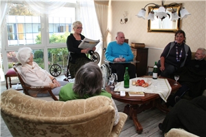 Das Foto zeigt einige ältere Menschen im Kreis sitzend, denen eine Altenpflegerin etwas vorliest.