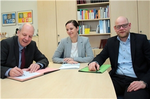 Das Foto zeigt zwei Männer und eine Frau an einem Tisch, der Mann links unterschreibt einen Vertrag.