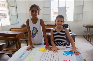 Das Foto zeigt zwei syrische Flüchtlingskinder in einem lichtdurchfluteten Raum, die stolz ihr mit bunten Händen gestaltetes Plakat zeigen.