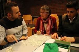 Das Foto zeigt zwei junge Flüchtlinge mit einer Frau, die an einem Tisch Scrabble spielen und sich unterhalten.