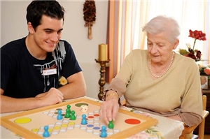 Das Foto zeigt einen jungen Altenpflege, der mit einer älteren Frau am Tisch sitzt.