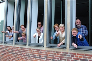 040-2018_ CKD 2025 Mitglieder der Projektgruppe gucken aus mehreren Fenstern eines Konferenzraumes und zeigen mit dem Finger nach vorne.