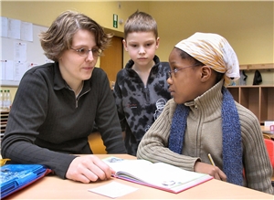 Das Foto zeigt zwei Grundschulkinder mit einer erwachsenen Frau an einem Schultisch.
