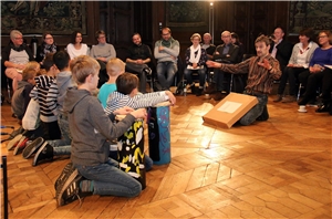 Das Foto zeigt eine Gruppe von Grundschülern seitlich von hinten, die vor einem im Kreis sitzenden Publikum trommeln.