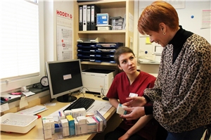 Das Foto zeigt eine junge Frau an einem Schreibtisch sitzend, die von einer stehenden Frau Medikamente erklärt bekommt.