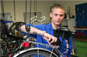 Das Foto zeigt einen jungen Erwachsenen in blauer Arbeitskleidung, der an einem aufgehängten Fahrrad schraubt.