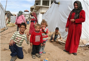 Drei kleine Kinder stehen auf trockenem Boden und schauen in die Kamera. Das Flüchtlingslager besteht aus notdürftig errichteten Unterkünften. Zelthütten aus weißer Plane sind mit Steinen befestigt.