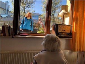 Das Foto zeigt eine alte Frau in einem Zimmer und eine Angehörige vor dem Fenster.
