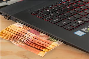 Das Symbolfoto zeigt einen Ausschnitt aus der Tastatur eines Laptops mit Geldscheinen, die aus dem DVD-Schlitz herausschauen.