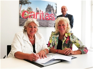 Zwei Frauen unterschreiben mit einem Lächeln den Stiftungsvertrag, im Hintergrund steht Diözesancaritasdirektor Heinz-Josef Kessmann