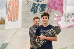 Ein Junge mit Skateboard ist an einer Halfpipe, hinter ihm steht ein Erwachsener Mann als Begleitung