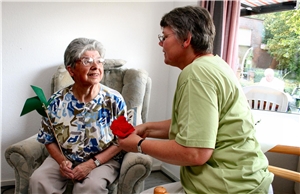 Das Foto zeigt eine Frau, die mit einer älteren, im Sessel sitzenden Frau spricht.