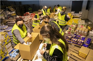 Das Foto zeigt Menschen in Warnwesten an einem langen Tisch, die Lebensmittel in Kartons packen.
