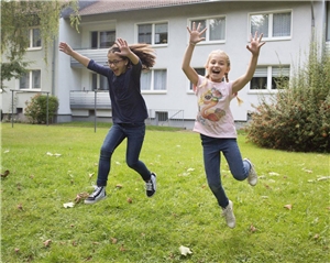 Das Foto zeigt zwei Mädchen, die auf dem Rasen vor einem Wohngebäude freudig in die Luft hüpfen.
