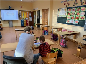 Das Foto zeigt eine Frau, die mit einem Jungen in einem Klassenraum sitzt, von hinten.