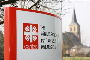 Das Foto zeigt eine Stele mit dem Logo des Caritasverbandes Geldern-Kevelaer und im Hintergrund leicht verschwommen einen Kirchturm.