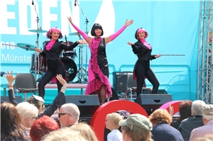 Drei Frauen in pink-schwarzen Kostümen tanzen auf einer Bühne vor Publikum.