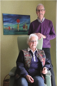 Das Foto zeigt ein älteres Paar. Die Frau sitzt im Sessel, ihr Mann steht hinter ihr.