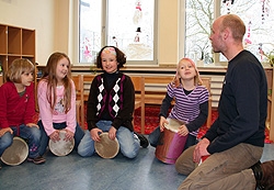 Vier Kinder sitzen mit dem Lehrer  auf dem Fußboden und musizieren mit ihren Trommeln