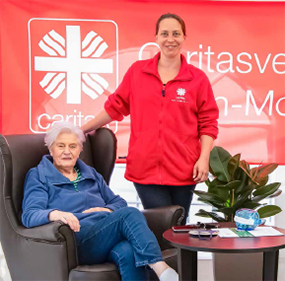 Eine Seniorin sitzt in Ledersessel in einer Sitzecke vor einem kleinen Tisch, neben ihr steht eine Caritas-Mitarbeiterin. Im Hintergrund ist ein rotes Caritas-Banner zu sehen.