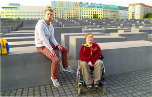 Ein Mann und eine Frau im Rollstuhl, die sich vor dem Holocaust-Mahnmal in Berlin befinden. Der Mann sitzt, neben einem bunten Rucksack, auf einem der Steine des Mahnmals.