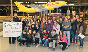 Eine Gruppe junger Spanierinnen und Spanier im Flughafen Paderborn-Lippstadt. Eine Frau hält ein Willkommens-Banner hoch, hinter der Gruppe steht ein aufblasbares Flugzeug.