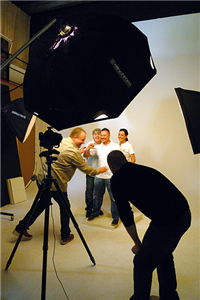 Drei Caritas-Mitarbeiter posieren in einem professionellen Fotostudio für ein Motiv der Kampagne Pflege-KRAFT gesucht!. Zwischen zwei Scheinwerfern u. der Kamera stehen der Fotograf u. sein Assistent.