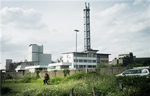 Der 'Stadtwerketurm' in Duisburg mit den umliegenden Gebäuden. Vor den Gebäuden erstreckt sich eine Wiese, auf der links ein alter Mann auf seinem Rollator sitzt und rechts ein silberner PKW steht.
