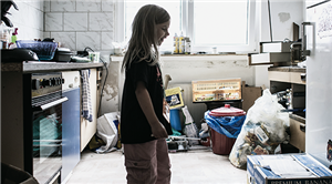 Ein Mädchen steht in einer unaufgeräumten Küche die auf der linken und rechten Seiten mit einigen Schränken und Geräten ausgestattet ist. Im hinteren Teil der Küche stehen einige Baumaterialien.